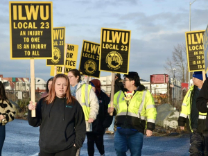 A group of union members and supporters march holding black and yellow ILWU Local 23 and Unfair Labor Practice signs.