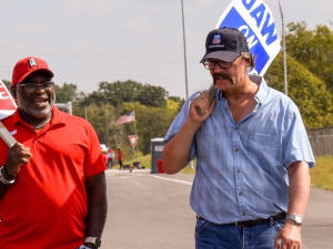 Two men, one Black and wearing red and one white wearing blue, walk together talking, each with a matching red or blue picket sign that says “UAW on Strike.”