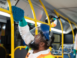 A worker cleaning a yellow pole with disinfectant on a NYC bus with another worker looking on. the workers are wearing orange safety vests, helmets, and transparent visors.