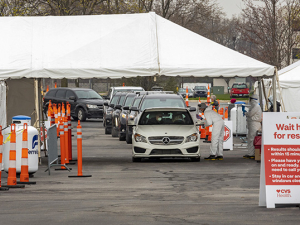 A line of cars coming through a coronavirus testing site in a parking lot. Site has tents.