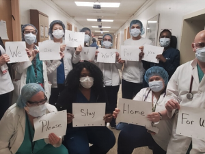 Masked health care workers in a hospital hold signs reading: "We... are... here... at... work... for.. you... Please... stay... home... for us."