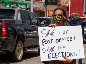 A masked protester holds a sign: "Save the Post Office! Save the Elections!" Behind the person, traffic and a "Coney Island" (diner) across the street