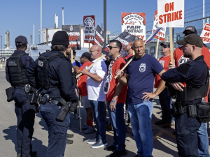 a group of picketers face three police officers. They are holding strike signs. A refinery truck is to the left.