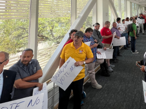 People in different colored union shirts or suits line both sides of a glass hallway. Many are men, some are Black and some are white. A few carry handmade signs urging a no vote on HB 1445.