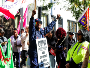 A Black worker in postal uniform, wearing reflective shades and a sandwich board sign that says "CUPW Toronto Local, We demand a fair contract," raises one fist in the air and a flag on a stick in the other hand. The flag is only partially visible but includes the word "Justice." This person is standing up on something we can't see, a step or box. Smiling people around them are holding various banners and signs, some wear safety yellow. Variety in race and gender. In sunlight dappled by shade of tree. 