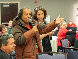 A person holding a mic stands speaking, gesturing with one arm, while others in the meeting listen intently. The group is diverse in race, gender, and age.