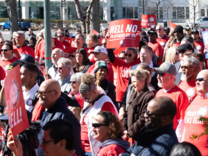 A large crowd of people in red "Fight Like Hell" shirts stand outside in bright sunlight. Some hold printed NALC signs with the slogans "Hell No to Attacks on Us" and "Fight Like Hell!"