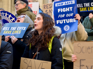 People, mostly white, rally outdoors on building steps. Printed signs say "Kill the cuts, save lives" and "UAW says: Fight for our future!" Handmade signs can't all be read but one says "Science belongs to everyone."