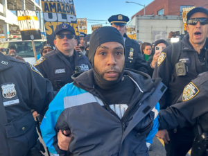 A man in a blue Amazon delivery driver jacket is flanked by bevy of cops, who are holding his arms and leading him away. More cops are behind him, and a crowd behind them. Printed Teamsters picket signs are visible saying "Amazon ULP strike" and "Amazon is unfair."