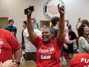 A Black woman in a red "Stand up UAW" T-shirt throws her hands in the air in celebration, phone in one hand, tears in her eyes. Other workers in matching shirts, and other people, stand nearby, all smiling, some clapping. In a room, looks like a union hall, with a logo on the wall that says "United we stand, divided we fall" around a handshake. 