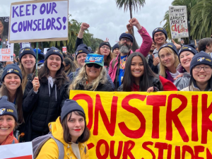 Smiling strikers pose outdoors with a bright yellow banner handpainted "On strike for our schools" in red. Most people visible in this photo appear to be white or Asian women. Many wear UESF hats. Other visible handmade signs say "Boo Su, give us a fair contract now!" "Keep our counselors" and "Invest in oiur largest asset: children." Palm trees are visible against gray sky. Some people raise fists or a victory/peace sign.