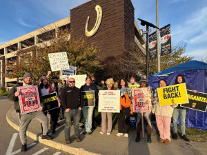A dozen or so workers stand on a corner in from of a parking garage with a giant horseshoe logo on it. They look determined. They carry a mix of hand-lettered and printed signs saying things like "Dealers on strike," "Organize," "Fight back," "Horseshoe Teamsters on strike," and "On strike only against Horseshoe Casino Greed, Teamsters Local 135." Behind them is a canopy that has been insulated with tarps closing some of the sides. Camp chairs and a propane tank are visible. Some wear knitted hats.