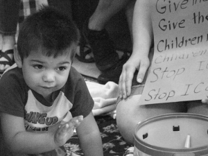 Child crouched in front of toys and a sign against ICE