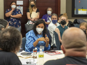 People are gathered around a table wearing masks, a woman with dark hair speaks and gestures in the middle.