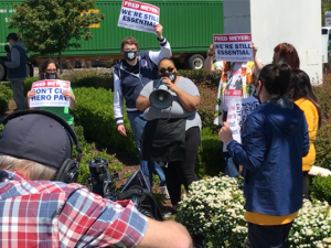 Woman with bullhorn surrounded at safe social distance by supporters with signs. Cameraman in foreground bottom left taking video.