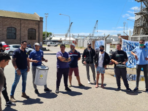 A group of workers, some holding drums, gathers in a circle outside a port.