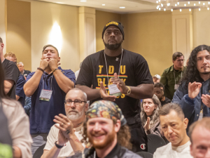 TDU convention attendees are shown sitting and standing, clapping for a speaker, in the hotel ballroom, under a chandelier.