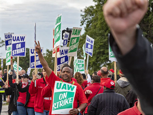 GM workers on a picket line, one work offscreen with arm raised, another centered with sign with arm raised.