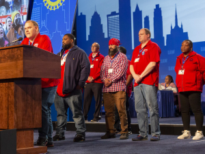 Autoworkers in red t-shirts stand on stage at a convention.