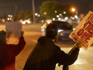 Workers hold signs at night--they are backlit by traffic and street lights but one sign is legible, saying ‘everyone tier one.’