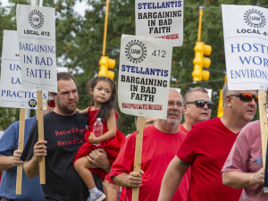 Auto workers march with signs that say “Stellantis Bargining in bad faith” One man is carrying a four-year old child.