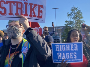 Amazon strikes hold signs saying strike, higher wages, stop racism. An Amazon facility is in the background