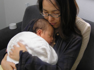 A seated woman with glasses in black holds a newborn baby wrapped in a white blanket to her chest.