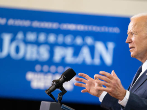 sideview of President Joe Biden speaking at a podium at Pittsburgh Carpenters Hall on March 31, 2021, with American Jobs Plan sign in background (white on blue)