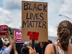 Crowd viewed from back. Sign in foreground, hand-lettered: "Black Lives Matter." Signs in background, printed: "Racism is a public health crisis" with the logo of National Nurses United
