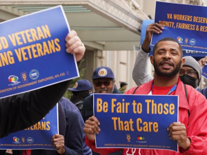 People (mainly Black men are visible) outside hold printed AFGE signs reading "Proud veterans serving veterans," "Be fair to those that care," and "VA workers deserve dignity, fairness, and respect."