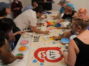 Several people work around a table adding colorful paints to a banner. Photo: ATF