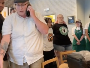 Starbucks workers in aprons surround a manager in a white shirt, the manager is leaving and talking on the phone