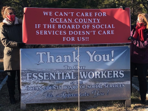 Two workers hold a banner reading “We Can't Care for Ocean County If the Board of Social Services Doesn’t Care About Us!” over the “Thank You to Our Essential Workers” sign that the Board had put up.