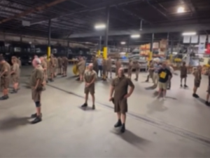 Indoors in a UPS warehouse, about 25 drivers are standing around the floor all with their backs to a manager with a yellow clipboard