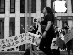 People march in front of an Apple store in New York City, holding signs and banners in English and Chinese. Their faces are blurred out in the photo due to fears of retaliation from the Chinese government against them and their families.