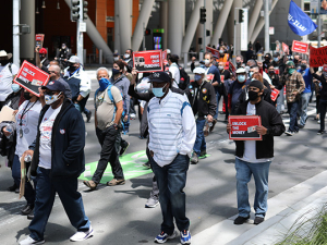 Masked people marching outside. Some have signs: "UNLOCK THE MONEY," "FREE OUR FUNDING," "AUSTERITY IS RACIST!"