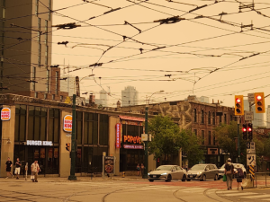 View of a city intersection. The sky, crisscrossed with power lines, is hazy grey and eerily tinted orange. Some people are out walking anyway.