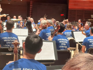 Orchestra musicians on a stage, apparently performing or rehearsing (conductor's baton is raised). Everyone, including the conductor, is wearing matching blue T-shirts that say "Philadelphia Musicians Union Local 77-274" on the back.