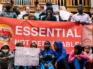 A diverse and serious crowd of people stands close together inside what looks like a church, in front of organ pipes, holding a big printed red banner that says "Essential not deportable" with the logo of Escucha Mi Voz. The person in the middle is speaking into a mic. Several kids sit in front and one holds a hand-lettered sign: "Free my grandma! Free her! I hate ICE!"