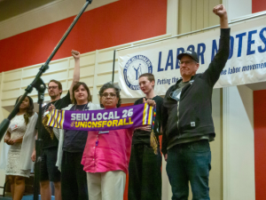 Six workers, of various races and genders, stand together on a ballroom stage with a big "Labor Notes" banner hanging behind them. Three are raising fists in the air, and one holds out a purple "SEIU Local 26, #UnionsForAll" scarf.