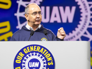 Shawn Fain, a white man with glasses, speaks at a podium. Behind him (and on the podium) is the UAW logo in white on a blue background. Yellow letters around it say (partly obscured) "Our Generation's Defining Moment."