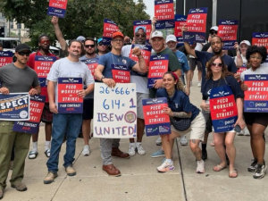 A group of workers pose for the camera on a city sidewalk. They are diverse in race, gender, and age. Many hold printed red and blue signs that say "PECO workers on strike!" One holds a big hand-drawn sign: "26+666+614=1. IBEW united!"