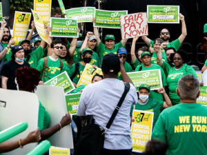 A crowd of 40 in green holds yellow and green signs saying “Fair Pay Now” and “Contract Now”