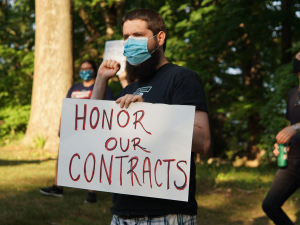 Masked person holding sign "Honor Our Contracts" in front of trees, other masked demonstrators