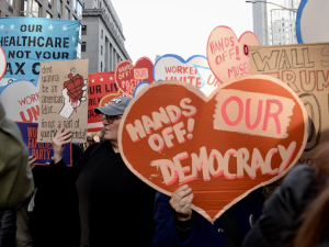 A heart-shaped sign saying “Hands Off Our Democracy” is carried in a crowd.