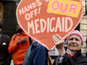 An older person holds up a handmade orange sign saying “Hands off our Medicaid”