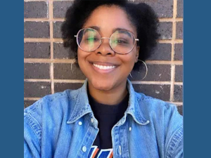 A smiling Black woman in round glasses and a jean jacket is shown