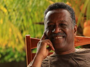 Chest-up photo of Dennis Serrette, a Black man, leaning back in a wooden rocking chair, resting his face gently on one hand, smiling warmly, bathed in warm evening light, with some greenery behind him.