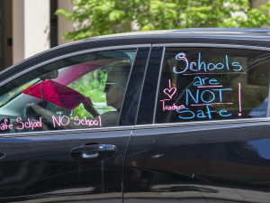 A woman drives a car with the words "Schools are not safe!" "I love teachers" and "People over profit" painted on the windows