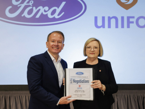 A man and a woman in suits stand holding a binder together in front of a screen that has Ford and Unifor logos projected on it.